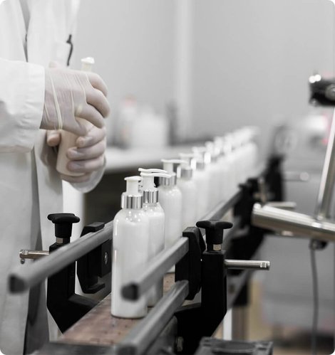 A technician in a lab coat and gloves tending white pump bottles on an APG production line